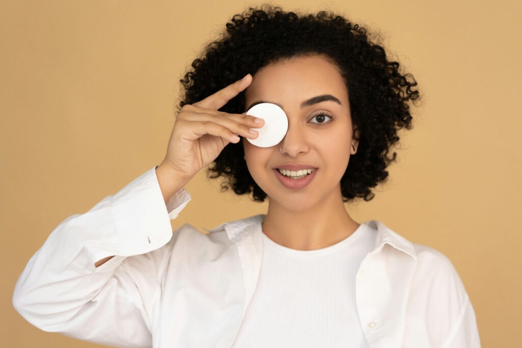 Smiling woman holding a cotton pad against her eye, enjoying skincare routine in a studio setting.